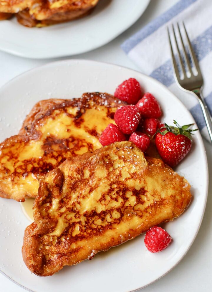 Two slices of challah bread french toast on a plate with raspberries, strawberries and maple syrup.