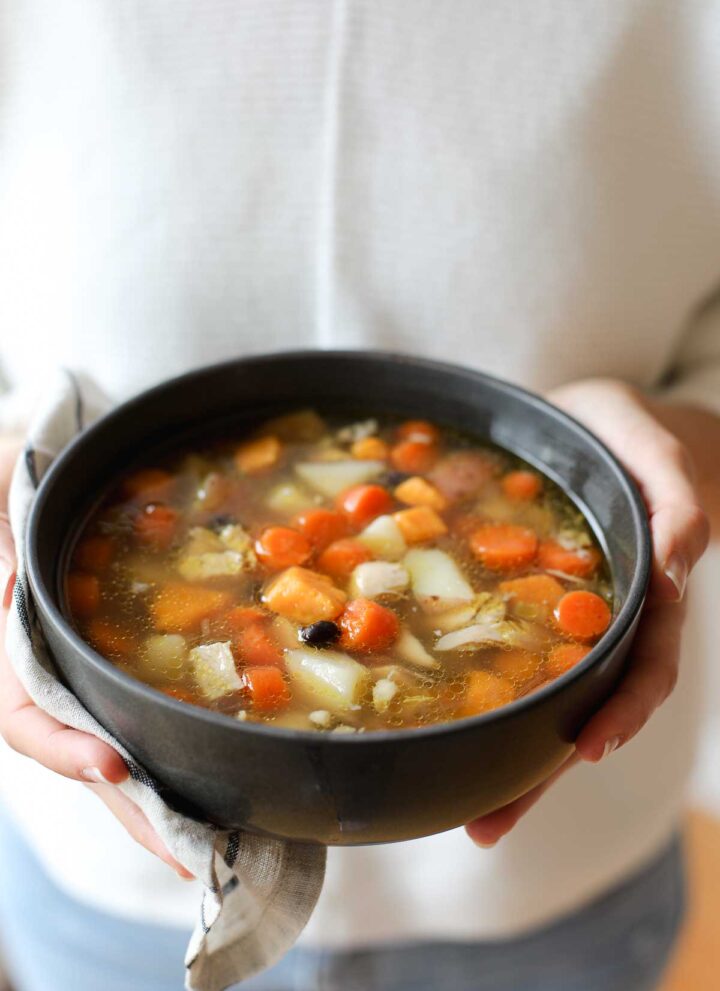Lauren holding a bowl of chicken and vegetable soup in a black bowl wearing a white sweater.