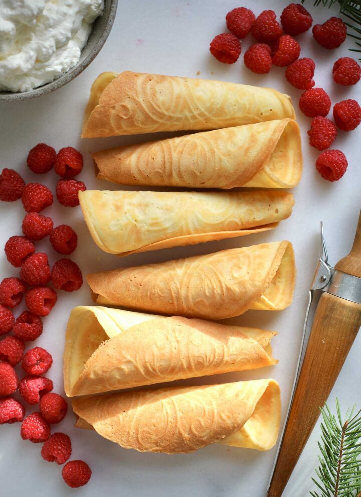 Krumkaker cones on a light background with a wooden dowel with a clip, raspberries and whipped cream on the side