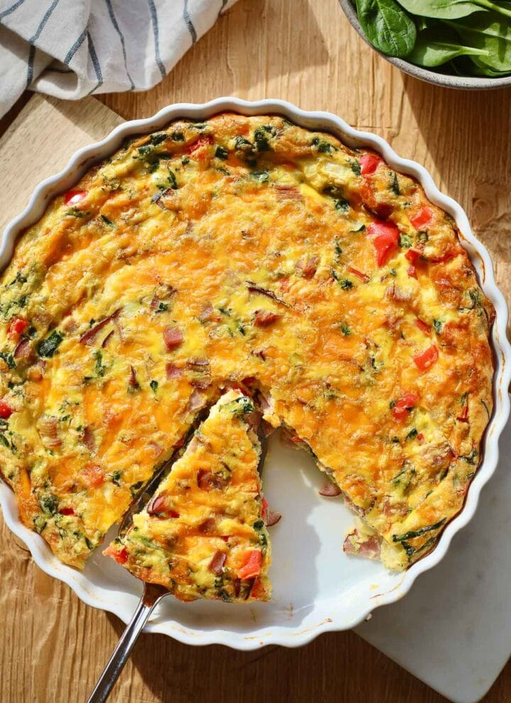 Ham and spinach frittata in a white quiche dish with a slice cut and being removed.