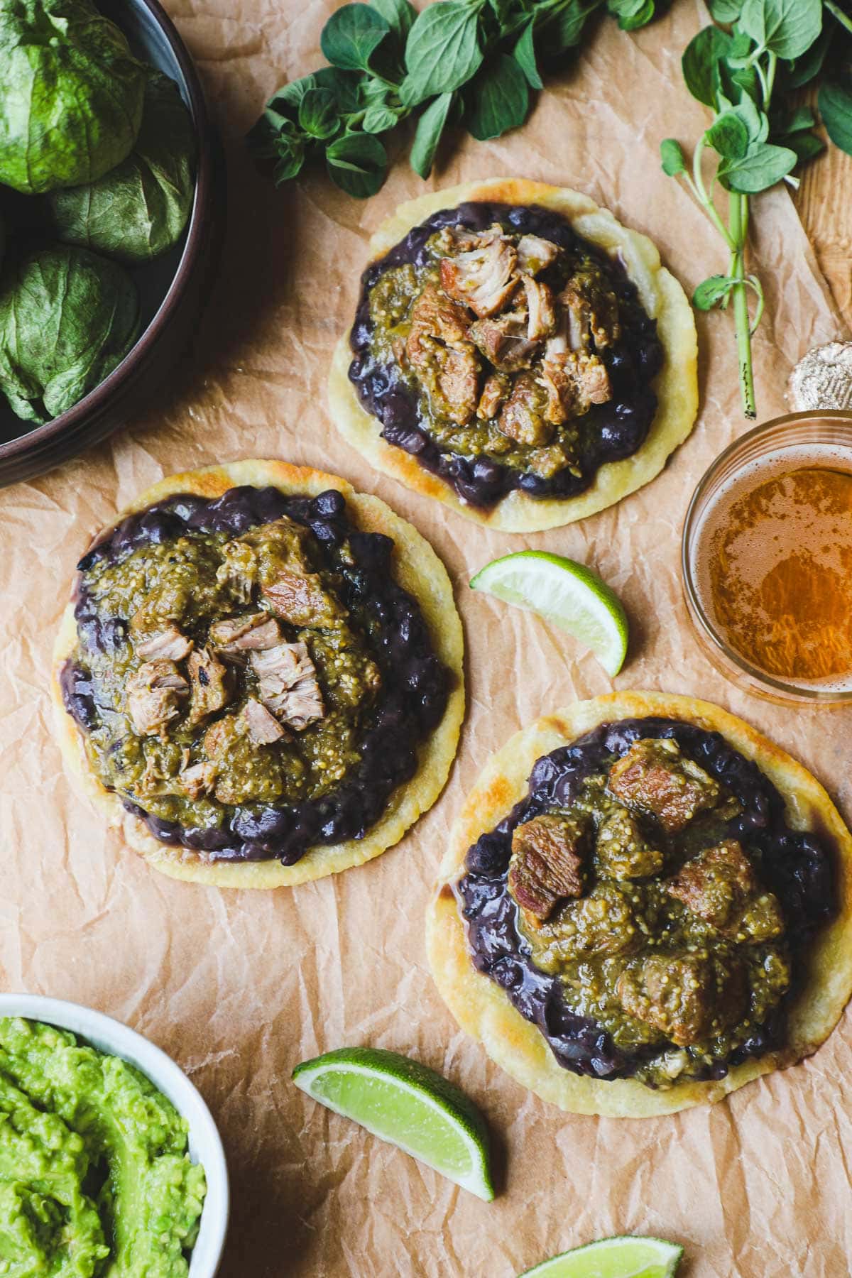 Three green chili pork tostadas with refried black beans arranged on brown parchment paper with limes, guacamole, extra tomatillos, and a bubbly beer.