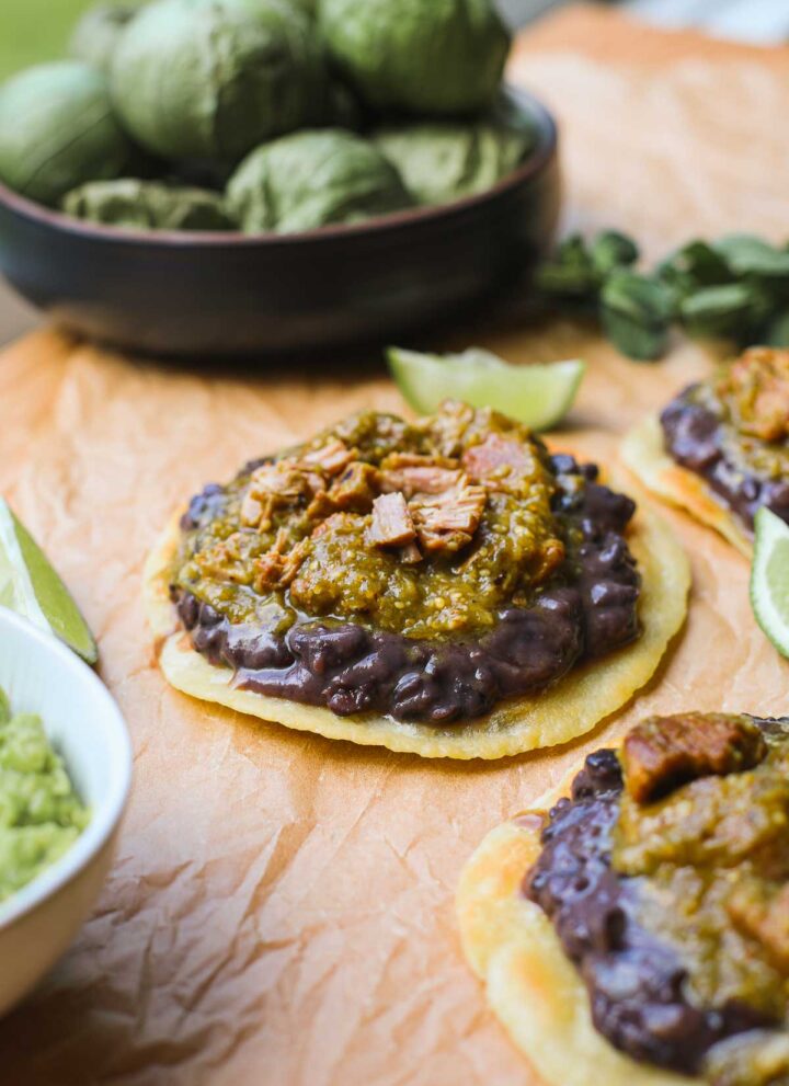 A crispy corn tortilla layered with refried black beans and green chili on a light brown background with more tostadas and tomatillos in the background.