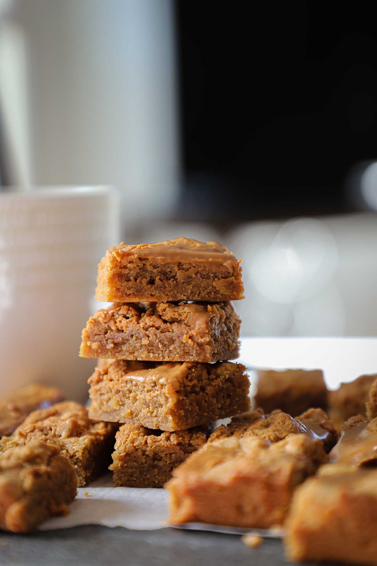 Four mini Biscoff Blondies stacked on top of each other next to a coffee mug surrounded by more Biscoff blondies.