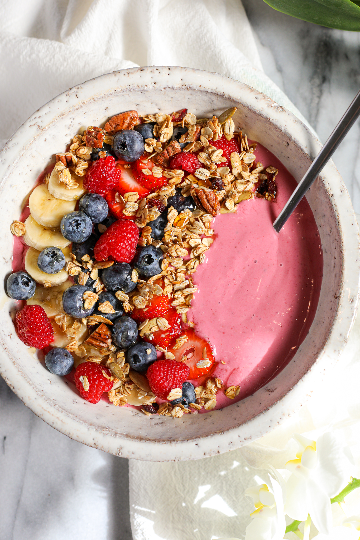 Bowl filled with brightly colored raspberry smoothie and topped with sliced bananas, sliced strawberries, blueberries, raspberries, granola, and honey.