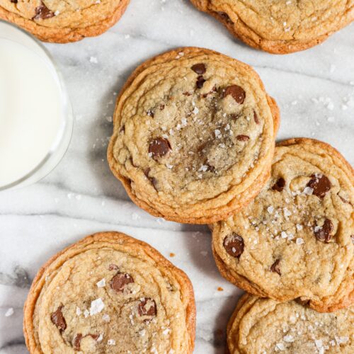 Several crispy chocolate chip cookies laying flat on a counter with a glass of milk.
