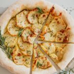 A potato and rosemary pizza on a white background with three slices cut surrounded by rosemary garnish.