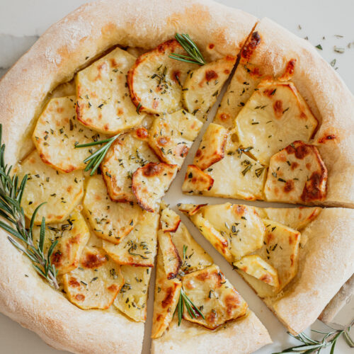 Round pizza with a thick chewy crust layered with thinly sliced potatoes and topped with rosemary with rosemary garnish on a white background.