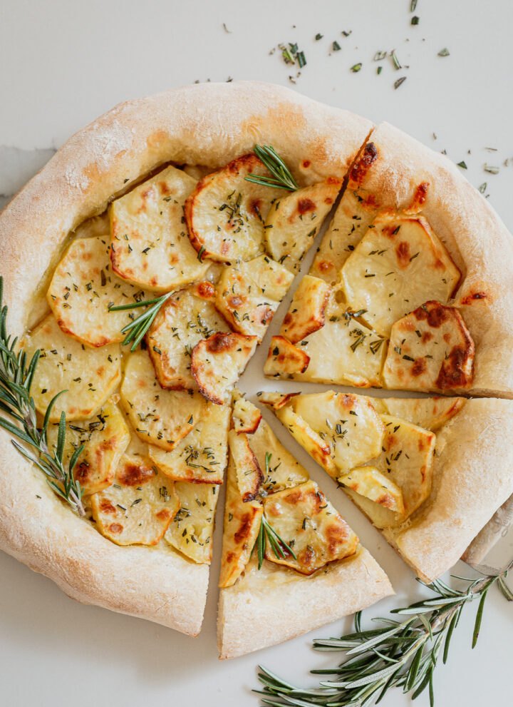 Round pizza with a thick chewy crust layered with thinly sliced potatoes and topped with rosemary with rosemary garnish on a white background.