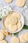 Soft drop sugar cookies on a cream colored plate with additional cookies and white flowers surrounding the plate.