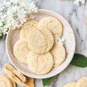 Soft drop sugar cookies on a cream colored plate with additional cookies and white flowers surrounding the plate.