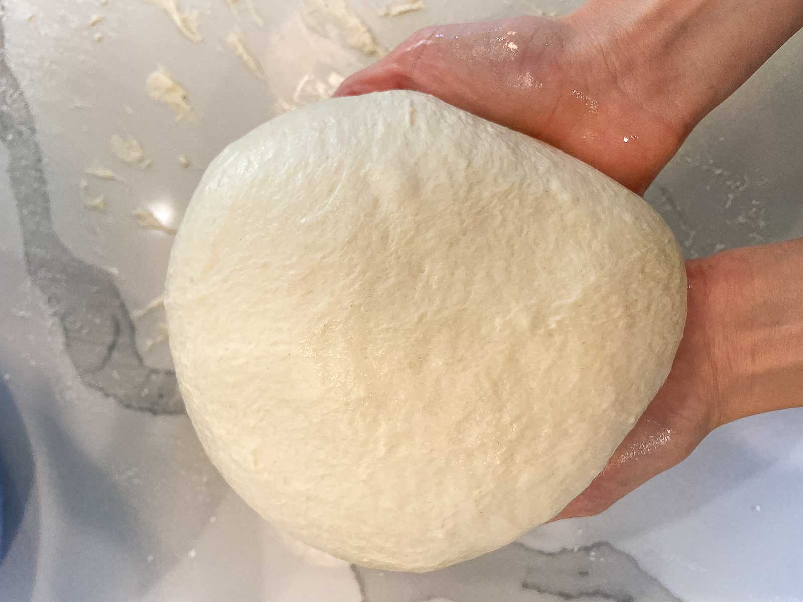 Round pizza dough being held in two hands on a light background.