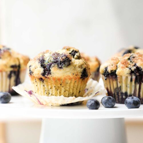 Blueberry Lemon Muffins on a cake stand with a white background decorated with blueberries and purple flowers.
