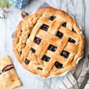 A Classic Blueberry Pie with a lattice braided pie crust surrounded by blueberry hand pies and blue flowers.