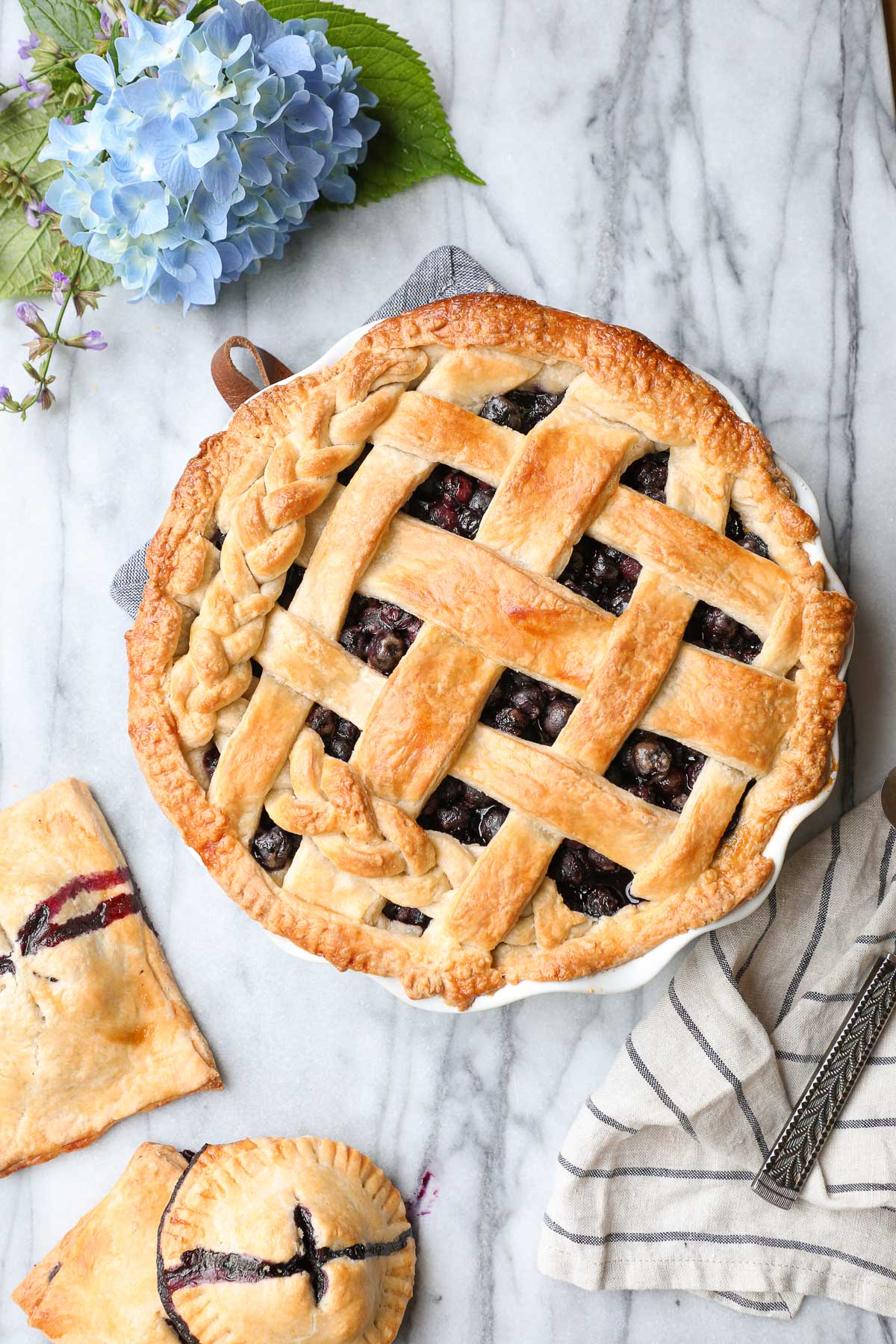 A Classic Blueberry Pie with a lattice braided pie crust surrounded by blueberry hand pies and blue flowers.
