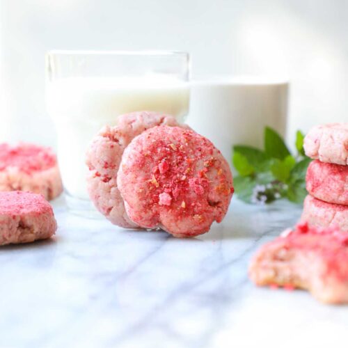 Two strawberry cheesecake cookies leaning against a glass of milk with a white background.