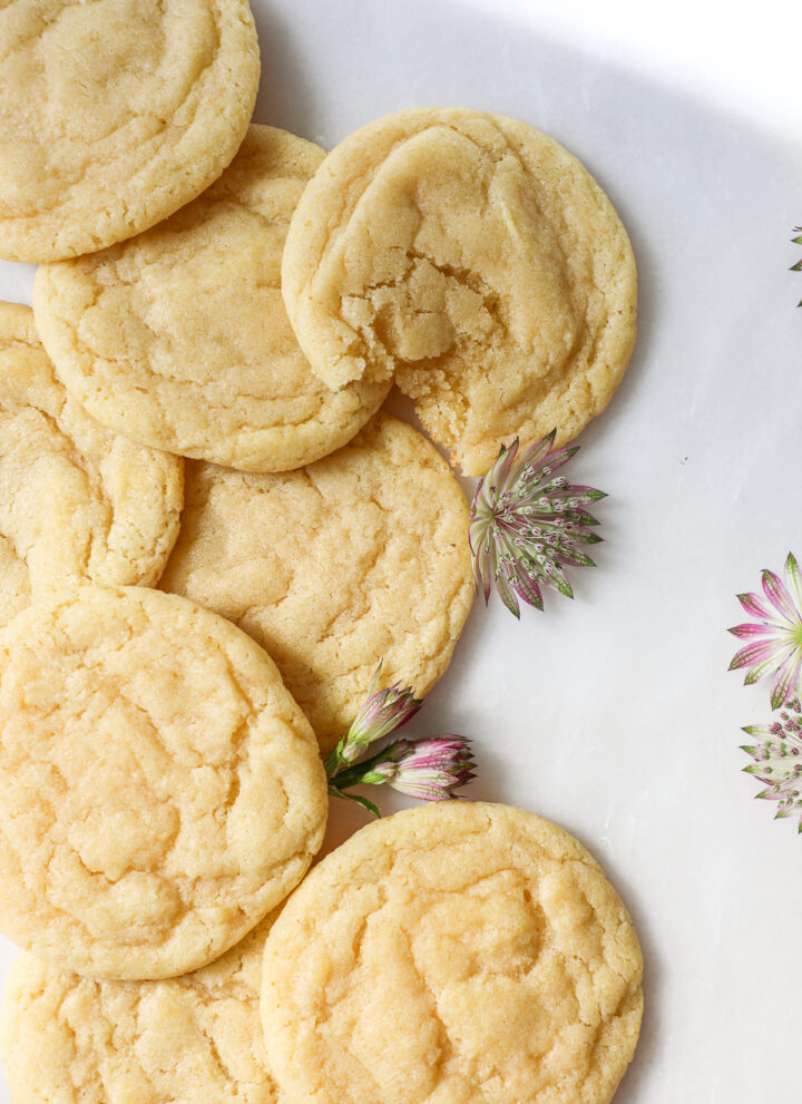 Soft drop sugar cookies overlapping each other laying on a flat white surface with decorative purple green and white flowers.