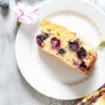 A slice of Lemon Blueberry Olive Oil Cake on a small white plate with a light marble background surrounded by powdered sugar, extra blueberries, and purple and white flowers.