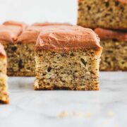 A square slice of Banana Cake covered in chocolate frosting on a light background with other cake pieces nearby.