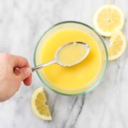 A hand holding a spoonful of Lemon Garlic White Wine Sauce with Butter over a bowl filled with the sauce on a light background.