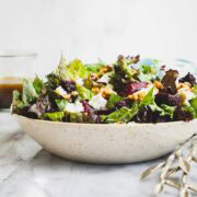 A Roasted Beet and Goat Cheese Salad in a tan speckled wide low bowl with a container of Balsamic Vinaigrette in the background.