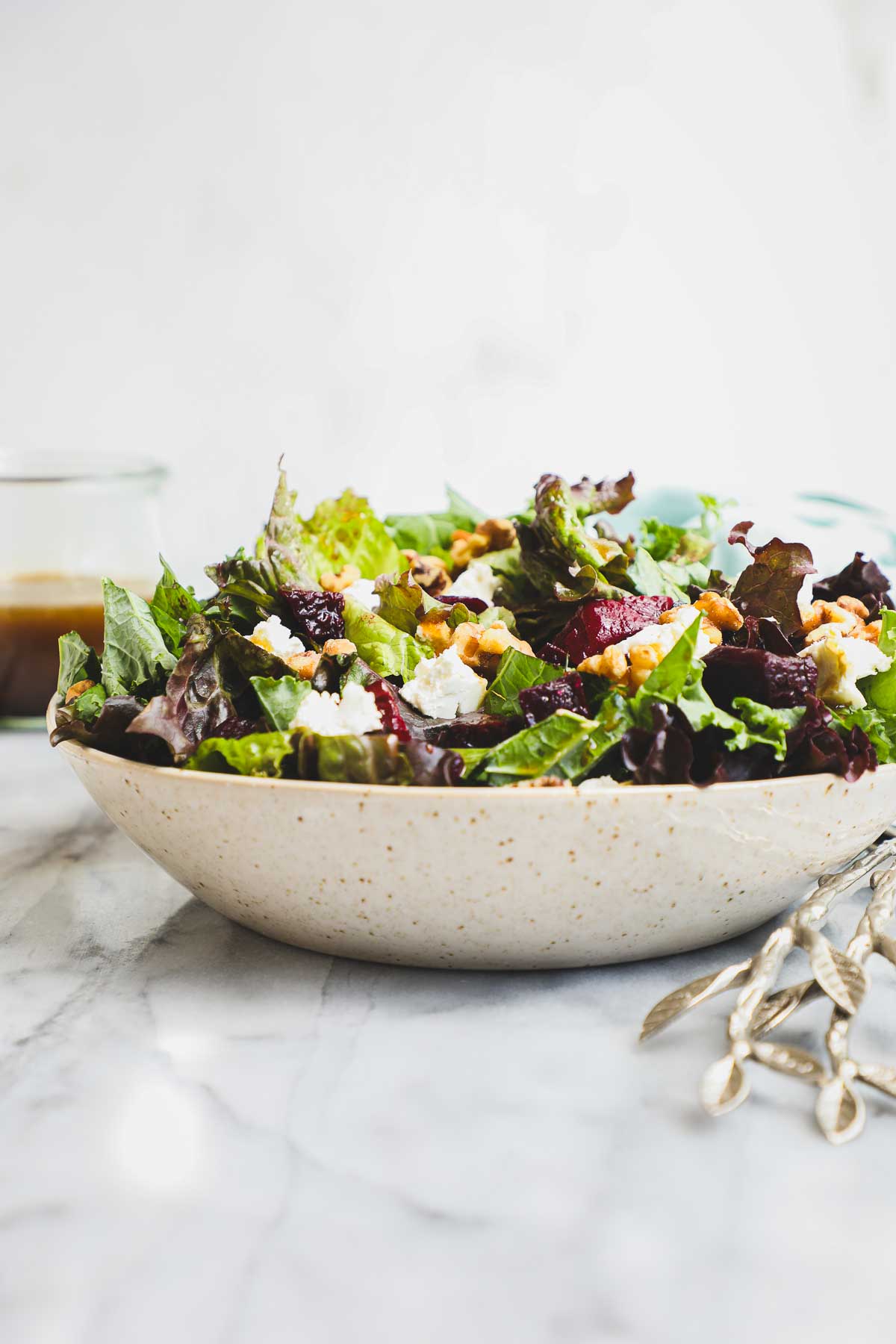 A Roasted Beet and Goat Cheese Salad in a tan speckled wide low bowl with a container of Balsamic Vinaigrette in the background.