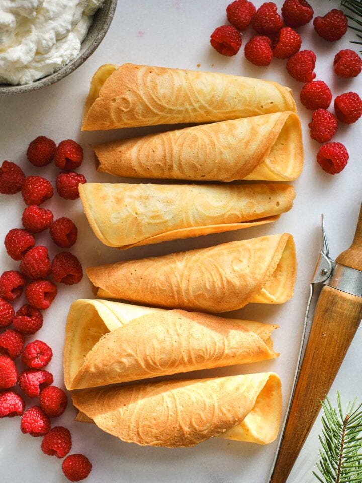 Norwegian Krumkaker waffle cookies lined up on a white board with raspberries and cream.