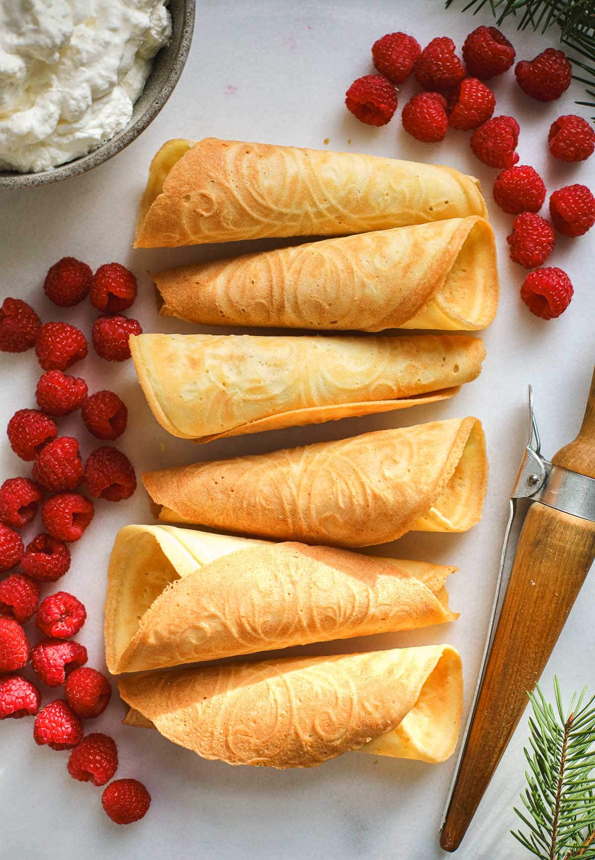Norwegian Krumkaker waffle cookies lined up on a white board with raspberries and cream.