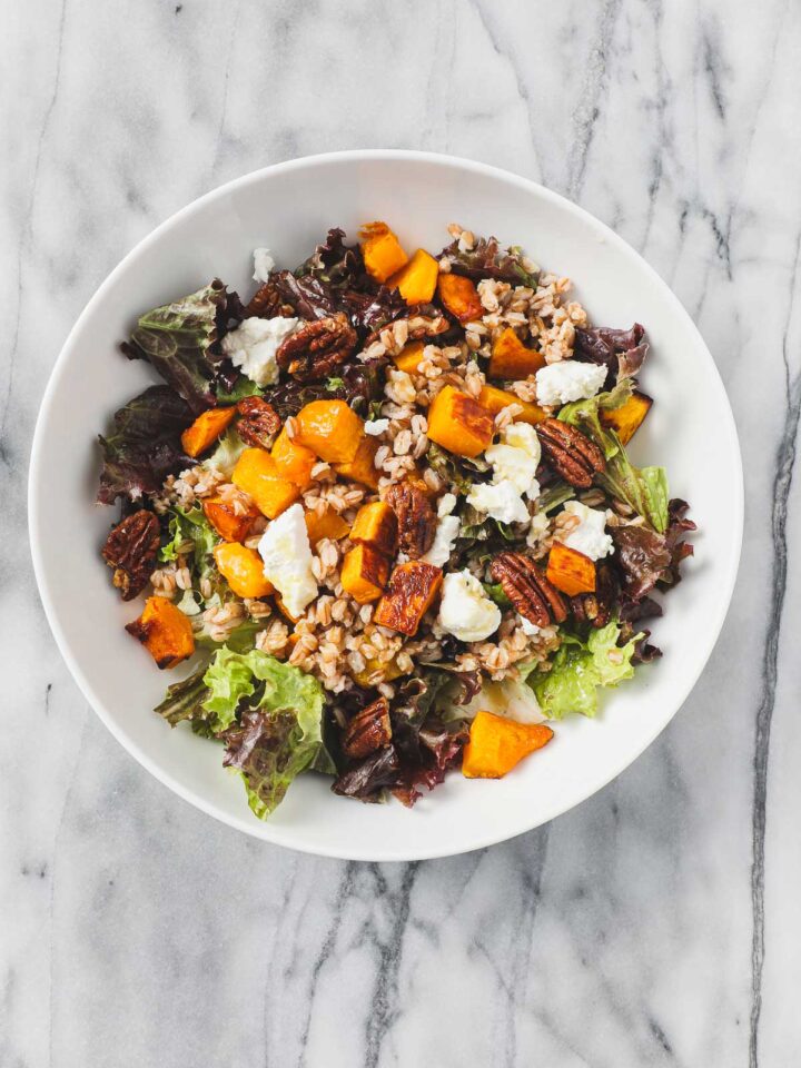 Butternut Squash Salad with Maple Vinaigrette in a large white salad bowl including maple roasted butternut squash, farro, goat cheese, and toasted pecans.
