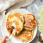 A white plate with four Fluffy Sourdough Banana Pancakes with butter and maples syrup and with one wedge being cut with a fork