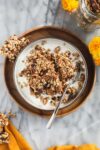 A bowl of yogurt topped with crunchy Sourdough Granola with yellow flowers and an orange yellow dish towel.