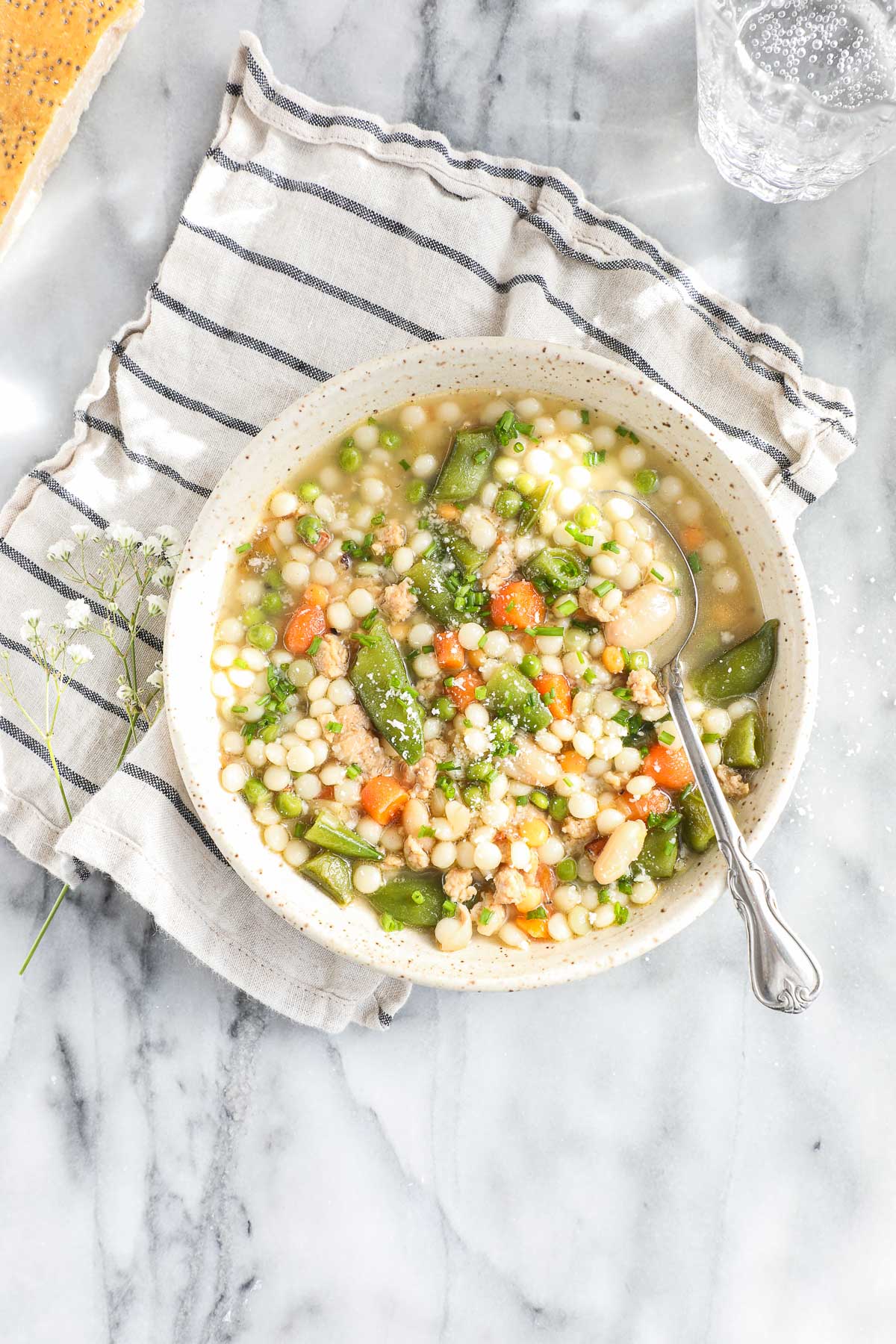 Spring Soup with Vegetables and Chicken in a light colored bowl with a spoon on a striped napkin with a sprig of white flours, a parmesan rind, and sparkling water in the background.