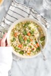 Spring Soup with Vegetables and Chicken in a light colored bowl with a striped napkin, a spoon, and Lauren's hand holding the bowl.