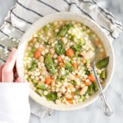 Spring Soup with Vegetables and Chicken in a light colored bowl with a striped napkin, a spoon, and Lauren's hand holding the bowl.