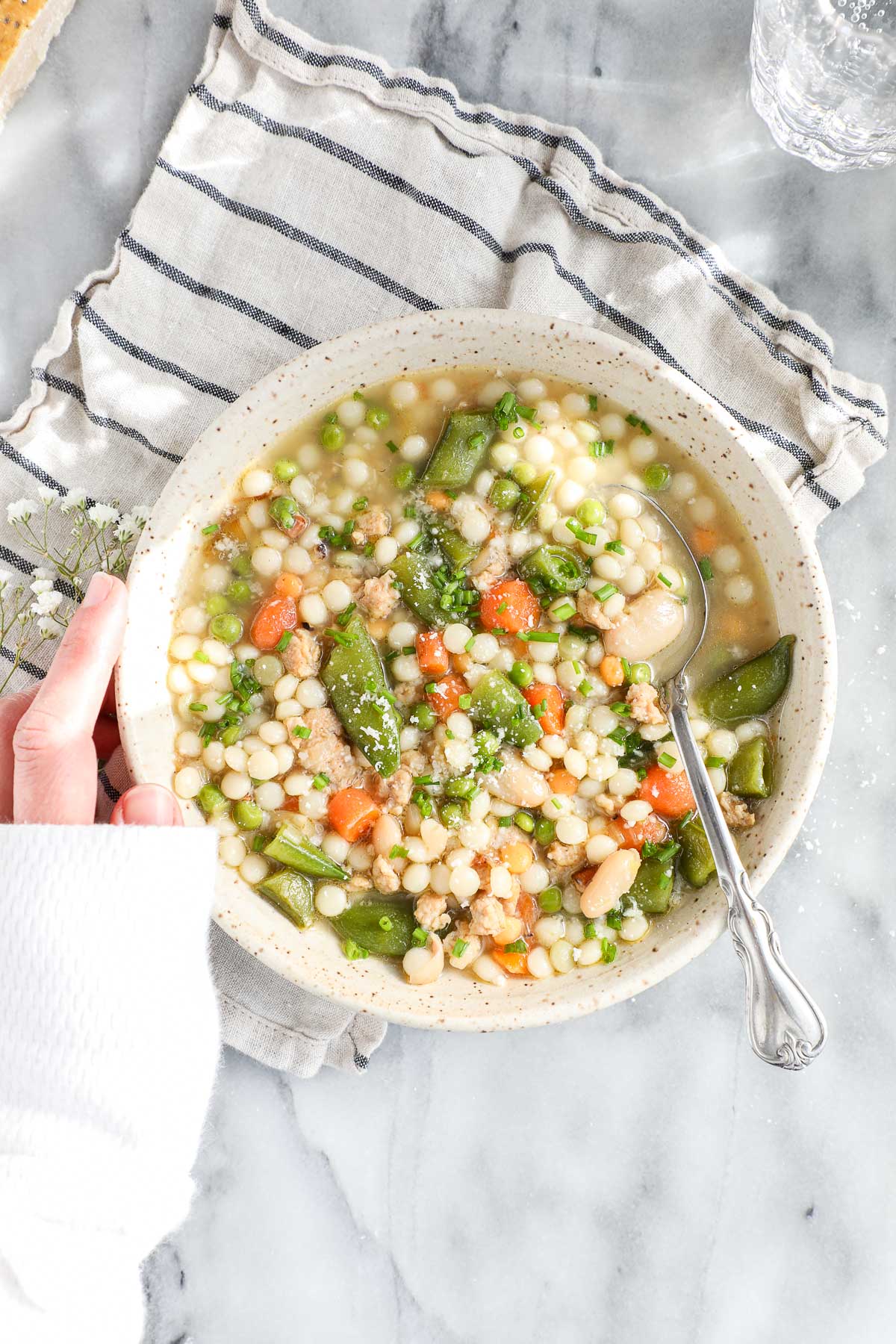 Spring Soup with Vegetables and Chicken in a light colored bowl with a striped napkin, a spoon, and Lauren's hand holding the bowl.