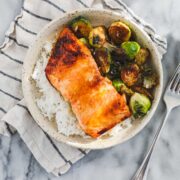A round shallow bowl with caramelized Hot Honey Salmon on top of white rice and pan roasted Brussel sprouts.
