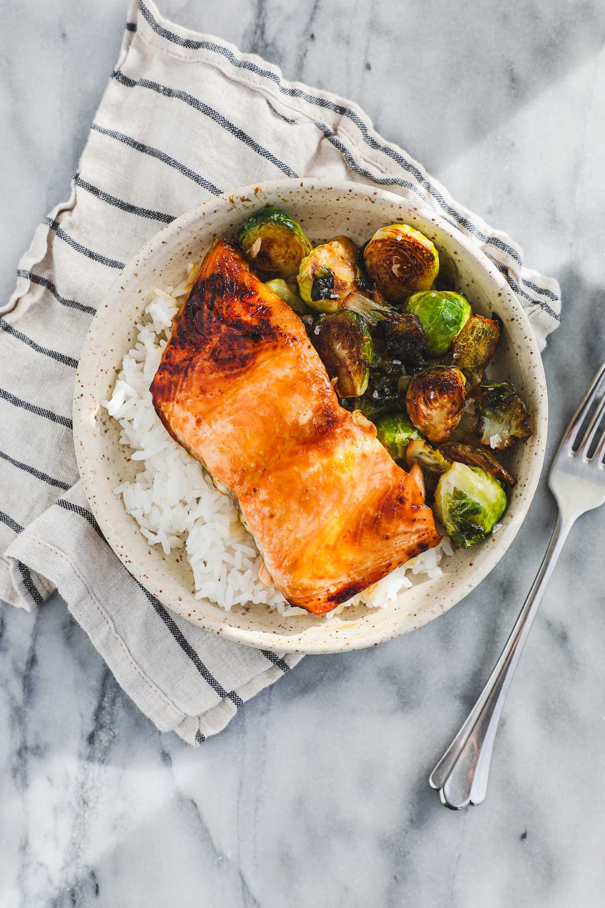 A round shallow bowl with caramelized Hot Honey Salmon on top of white rice and pan roasted Brussel sprouts.
