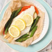 White fish layered on top of red bell peppers and zucchini with thinly sliced lemons on top in parchment paper that has been opened on a light blue plate.