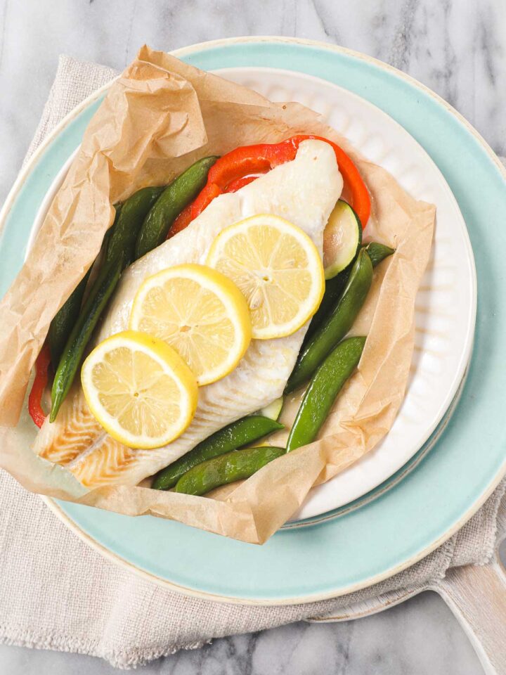 White fish layered on top of red bell peppers and zucchini with thinly sliced lemons on top in parchment paper that has been opened on a light blue plate.