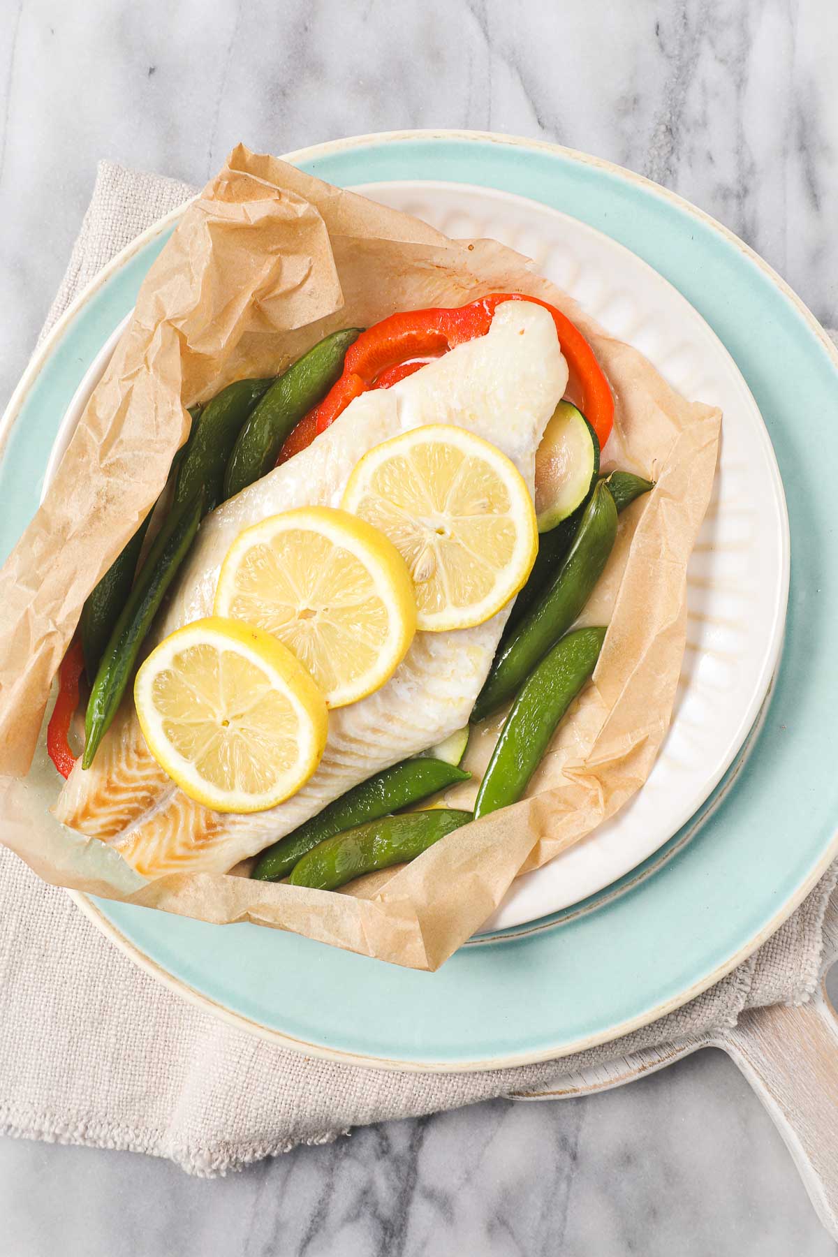 White fish layered on top of red bell peppers and zucchini with thinly sliced lemons on top in parchment paper that has been opened on a light blue plate.