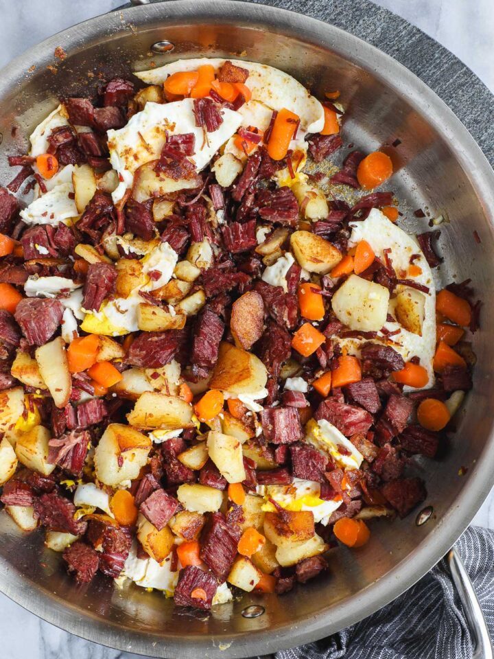 Corned Beef Hash in a large sauté pan with crispy corned beef, crispy potatoes, carrots, and pan fried eggs.