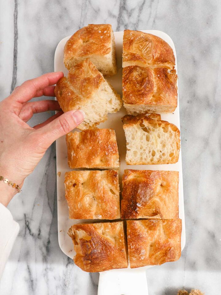 Loaf Pan Focaccia cut up into pieces and placed on a marble board with one piece turned on the side and Lauren picking up and holding another piece.