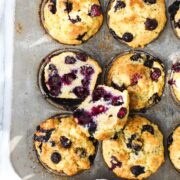 Olive Oil Blueberry Muffins in a muffin tin with one muffin cut in half to show the soft interior of the muffin filled with blueberries.