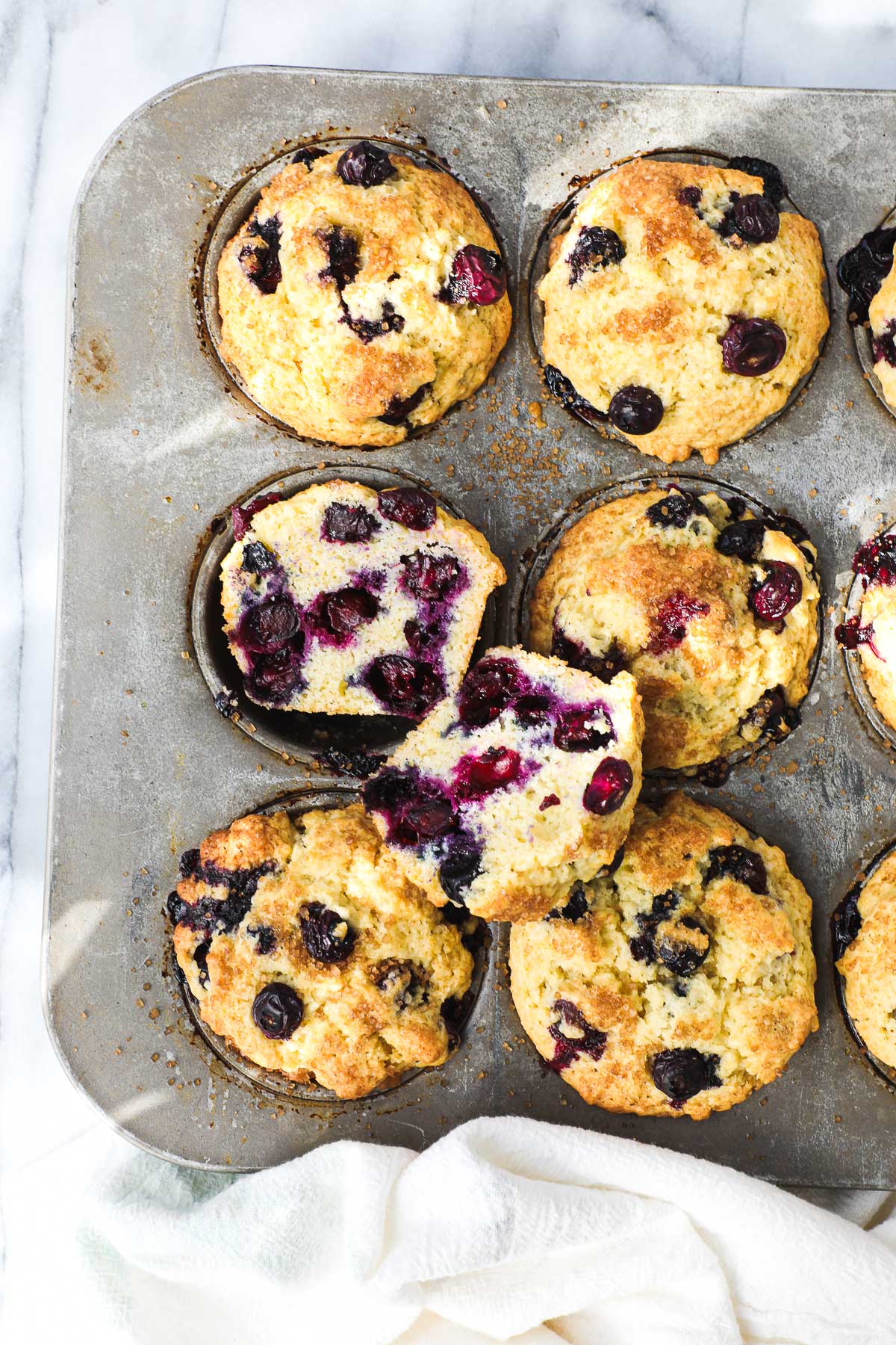 Olive Oil Blueberry Muffins in a muffin tin with one muffin cut in half to show the soft interior of the muffin filled with blueberries.