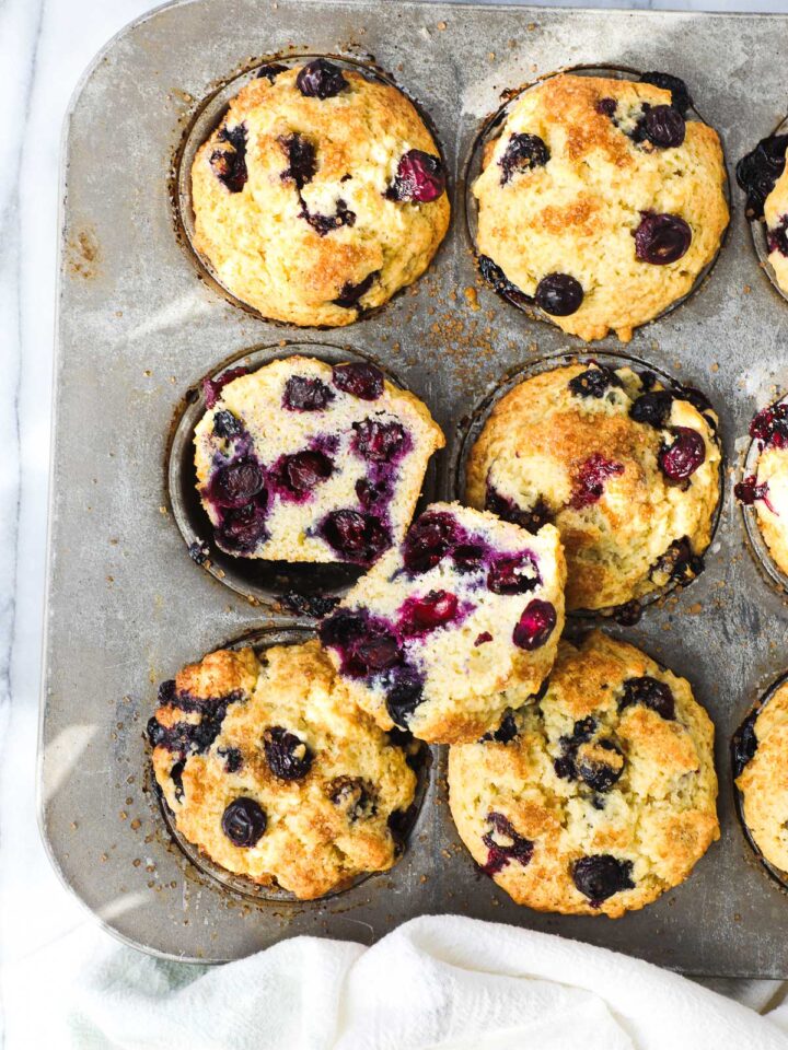 Olive Oil Blueberry Muffins in a muffin tin with one muffin cut in half to show the soft interior of the muffin filled with blueberries.
