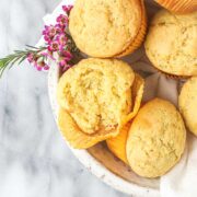 Several Olive Oil Muffins in a white serving bowl, with one muffin broken open to show a light fluffy interior.