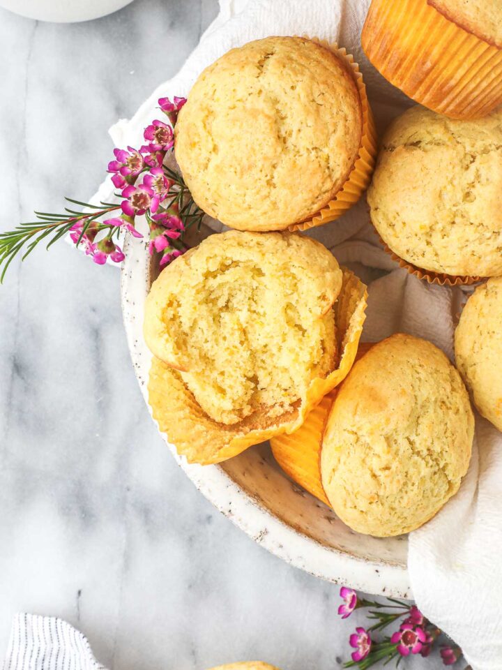 Several Olive Oil Muffins in a white serving bowl, with one muffin broken open to show a light fluffy interior.