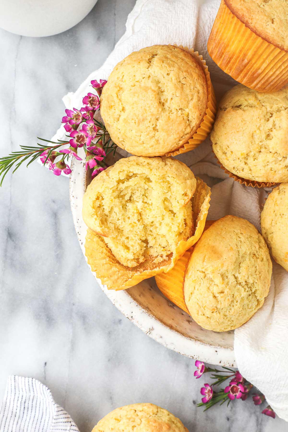 Several Olive Oil Muffins in a white serving bowl, with one muffin broken open to show a light fluffy interior.