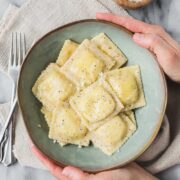 A blue green bowl with Ravioli Cacio e Pepe being held by Lauren on light background.
