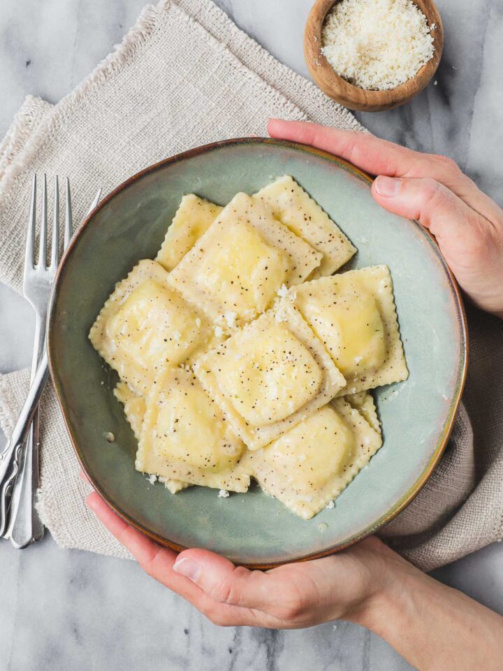 A blue green bowl with Ravioli Cacio e Pepe being held by Lauren on light background.