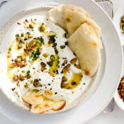 A shallow white bowl filled with thick and creamy lemony whipped feta topped with chopped pistachios, red pepper flakes, and minced parsley.